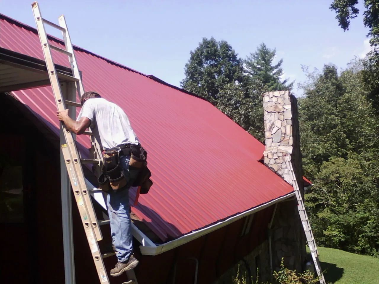 Roofer on a ladder beside a red metal roof and stone chimney — Soffit & Fascia Repair by Roofing Brothers in Gulf Shores